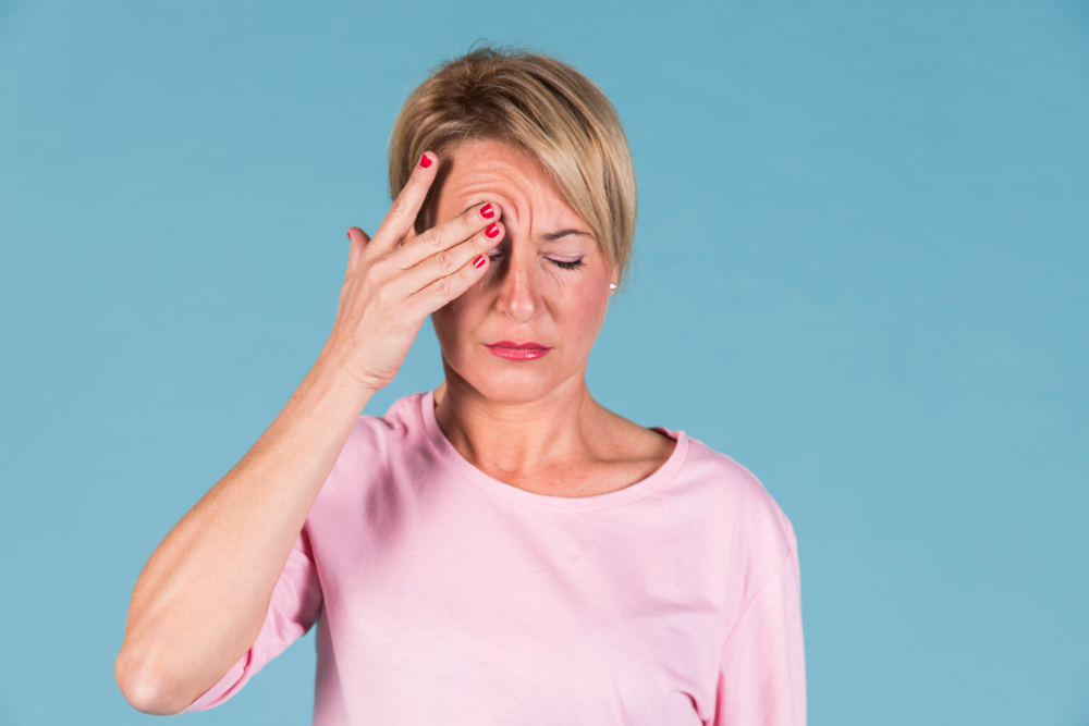 Une femme avec un pull rose, se tient la tête de douleur sur un fond bleu